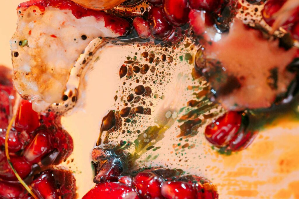 Abstract close up photograph of pomegranate seeds and various liquids on reflective surface. 