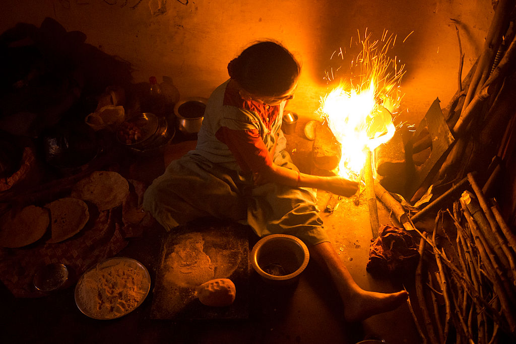 A woman wearing a red top and white apron sits in a dark room tending a fire, surrounded by plates and bowls holding food.