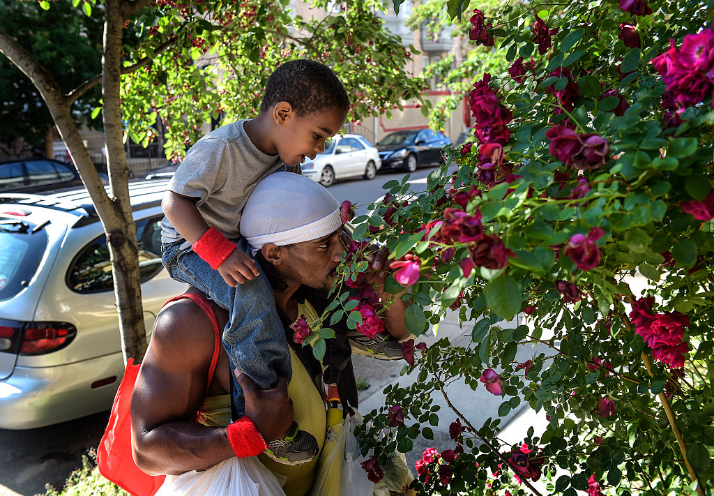 A man wearing a white durag and lime-green tank top props a young boy in a gray T-shirt and jeans on his shoulders while holding a pink rose from a rose bush to his nose.
