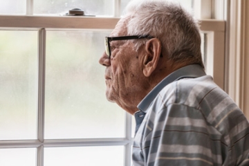 Older adult man feeling isolated and alone, staring out the window of his home.