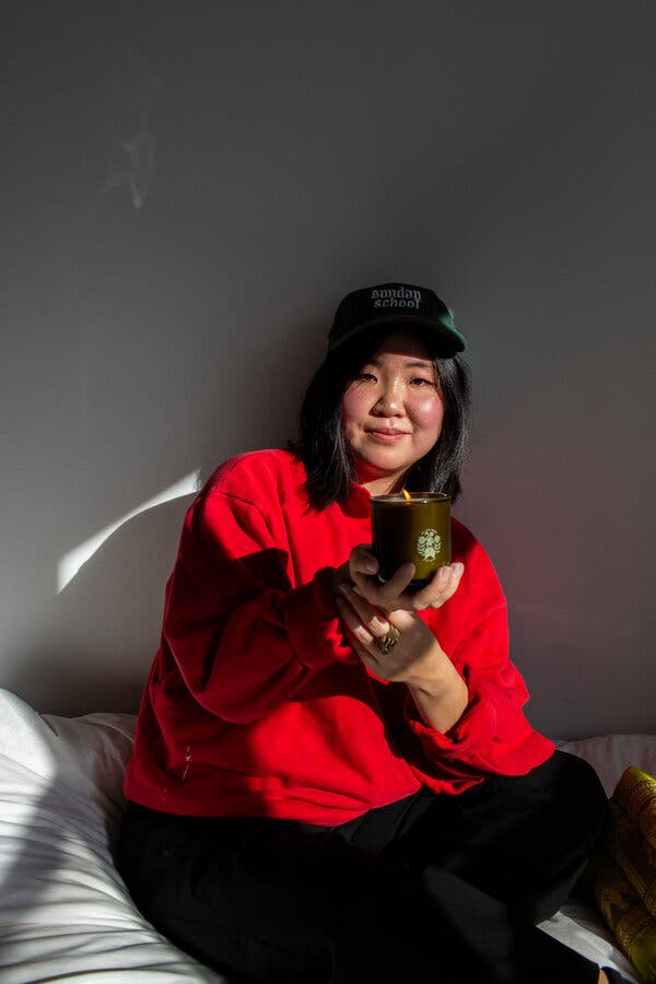 A person in a red sweater, with black hair and a baseball cap that says “Sunday School,” sits against a blank wall holding a lit candle.