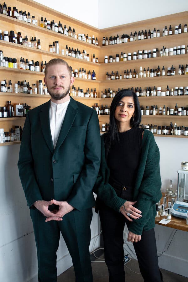 A couple, one in a suit and the other in black with a green cardigan, stand against shelves filled with many different kinds of vials.